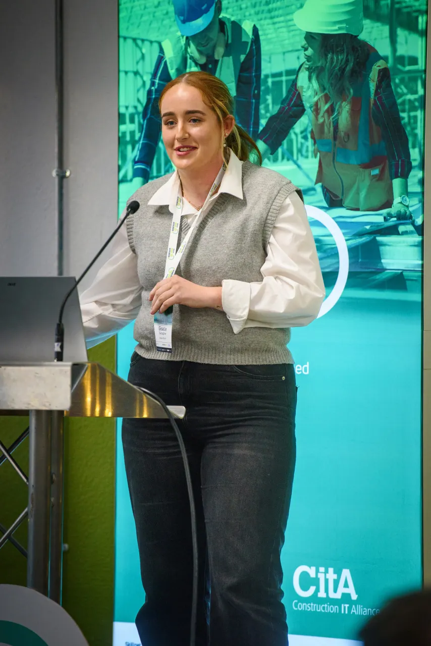 Person in white shirt and black pants standing at a microphone in front of turquoise backdrop.