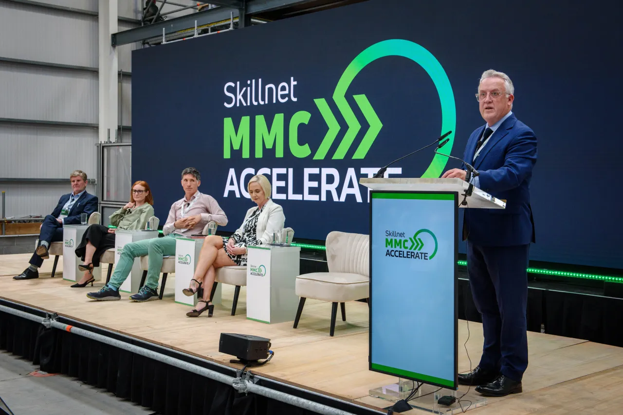 A speaker at a podium during the Subpact Accelerate event, with panelists seated on stage.