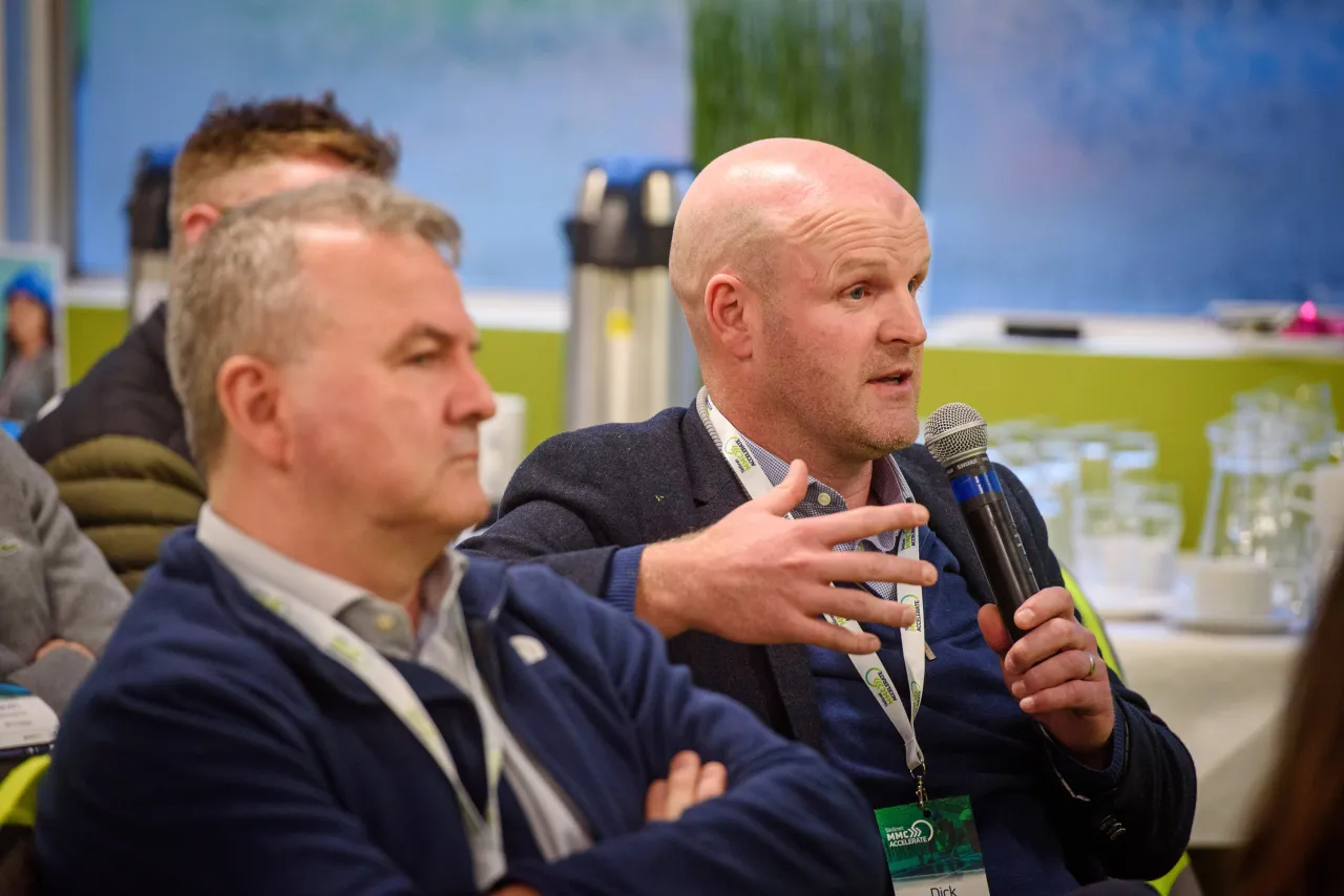 Two men in suits gesturing during a business event with colorful displays in background.