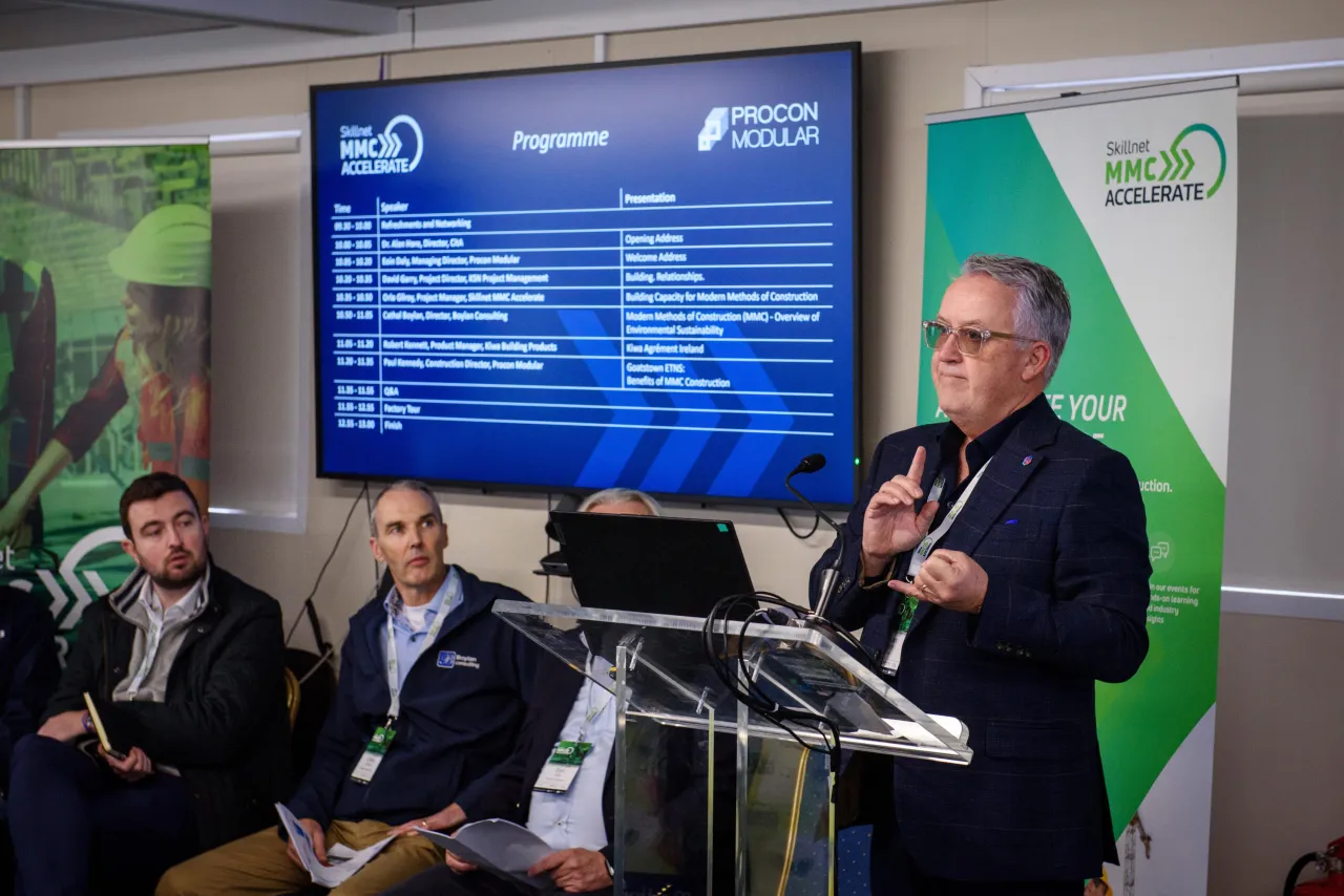 A person speaking at a podium during a conference with a digital screen and seated panelists in the background.