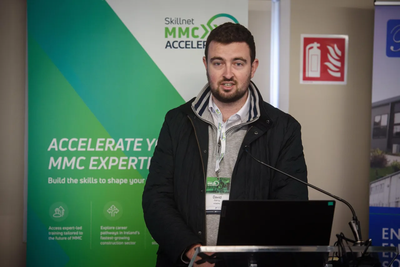 A person in a black jacket with an event badge standing at a podium with "Accelerate" signage in the background.