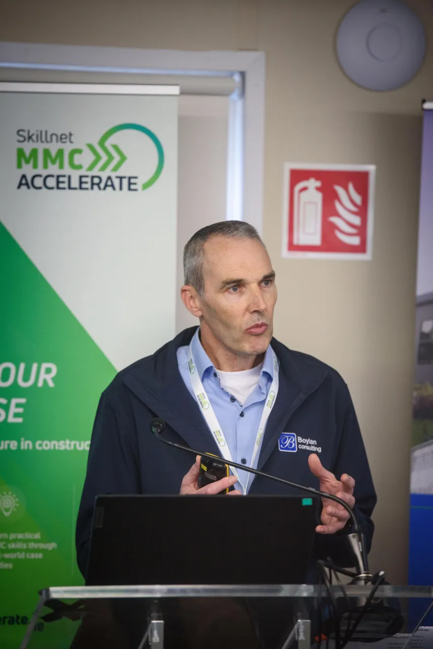 Person in a suit holding a microphone, speaking at a podium with a MISC ACCELERE sign.