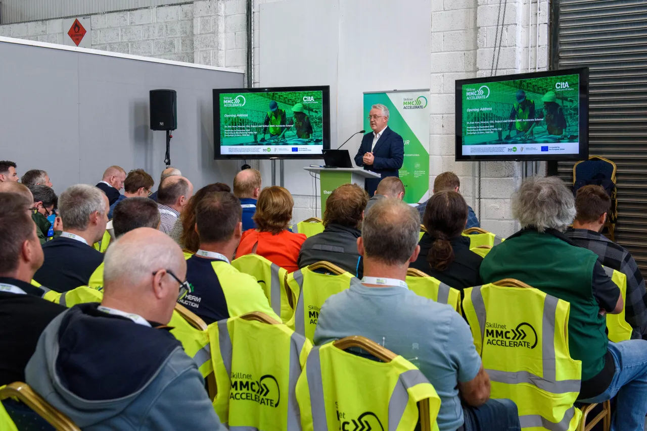 A person presenting to workers in safety vests at a construction event with screens showing data.
