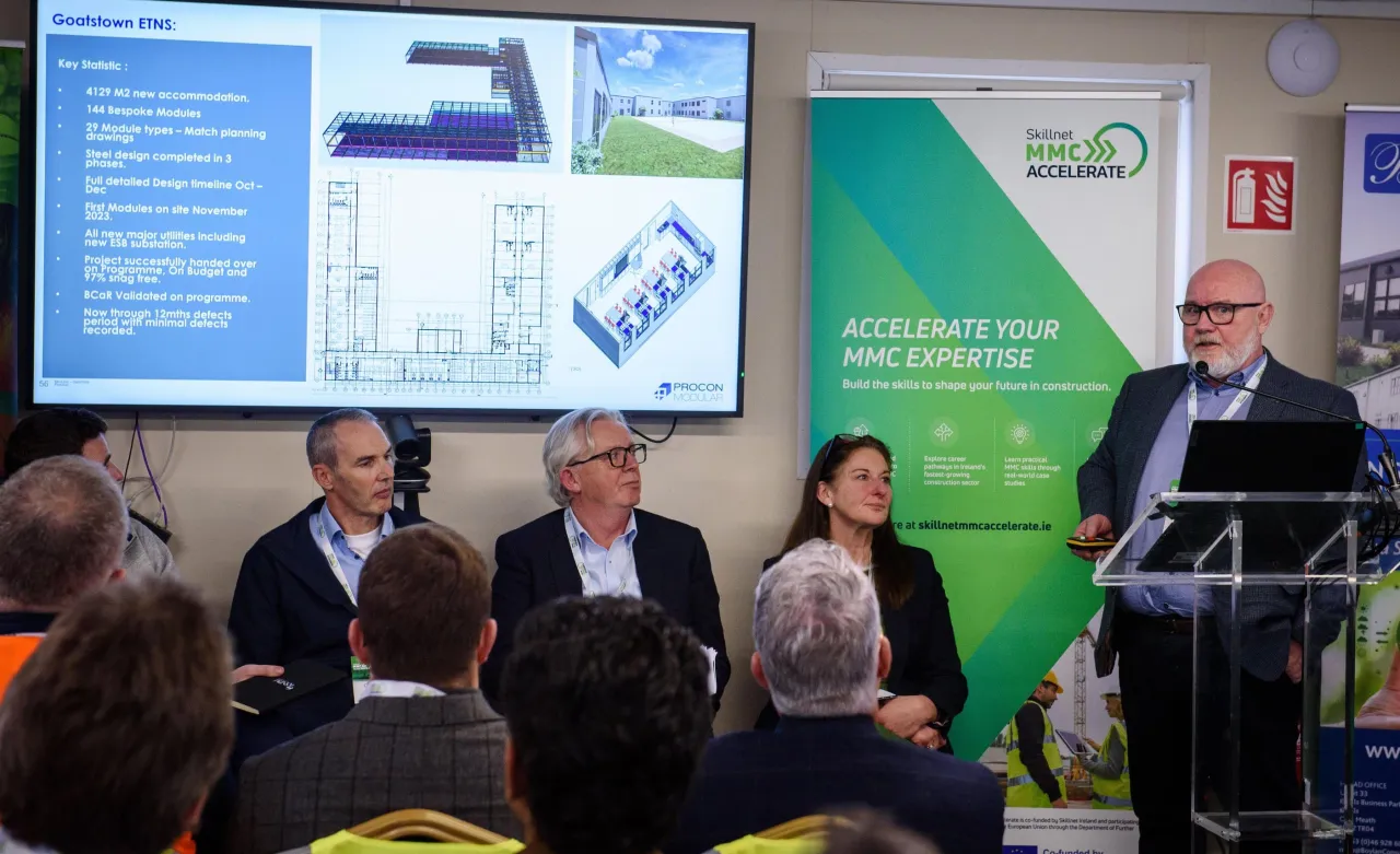 Speaker at podium with panel seated at table during a business presentation with projector screen and banner.