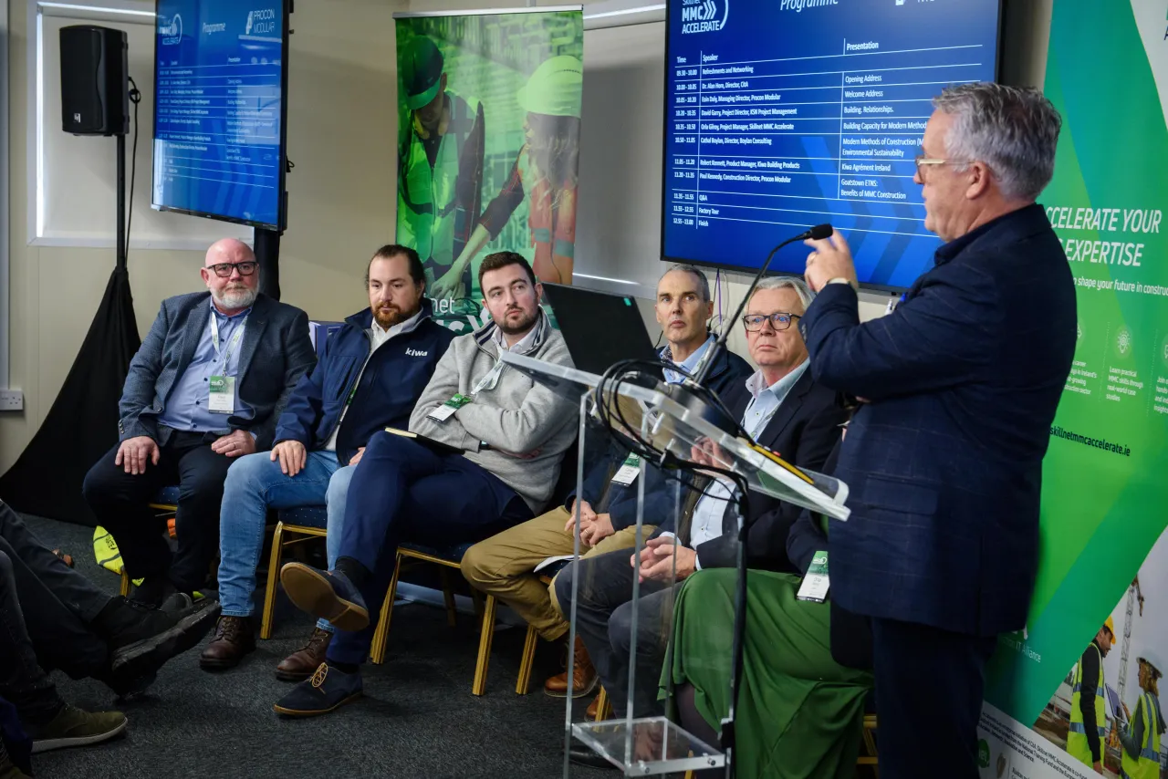Panel discussion with speakers seated on a stage, one person standing at a podium addressing the audience.