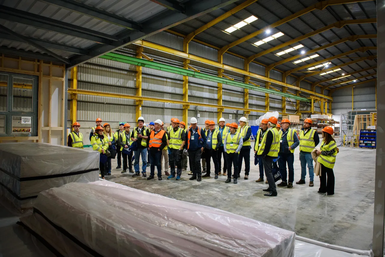 Group of workers in safety vests and helmets standing in a large industrial warehouse with stacked materials.