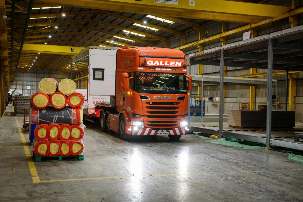 Red Scania truck parked inside industrial warehouse with stacked wooden barrels nearby.