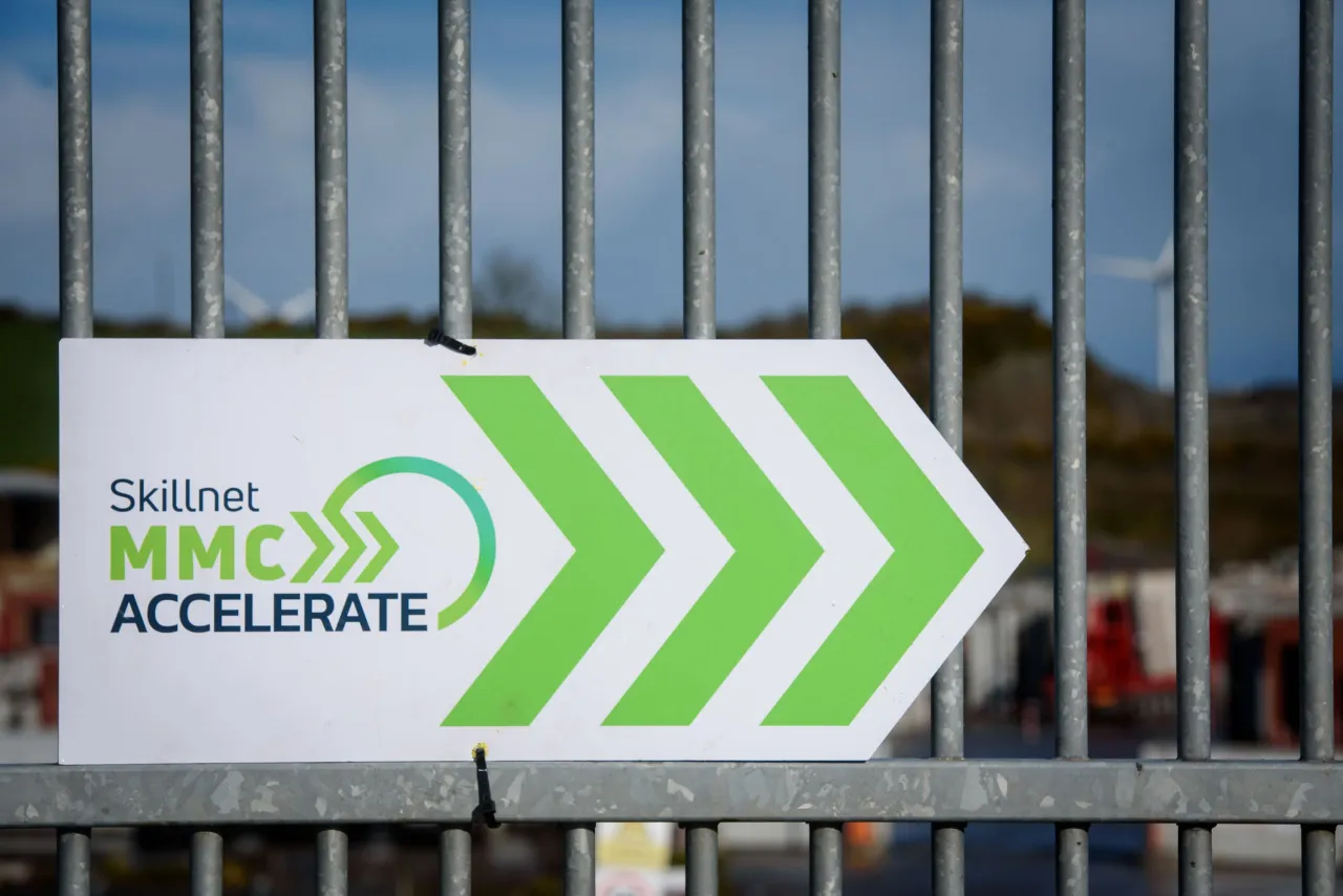 White arrow sign with green chevrons reading "Skillnet MMC Accelerate" mounted on a metal fence.