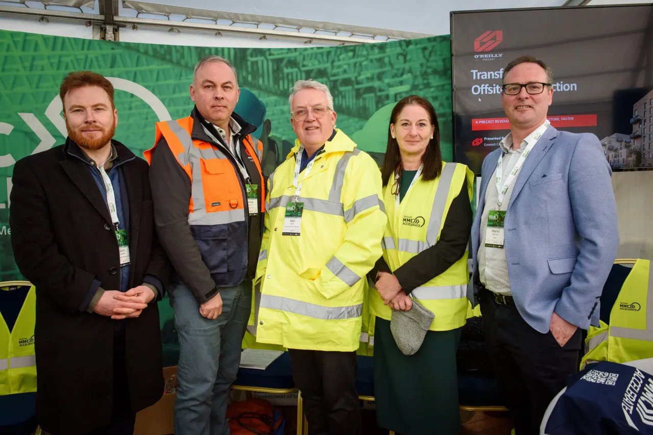 Five people posing together outdoors, two wearing high-visibility safety vests, at an event with green banners.