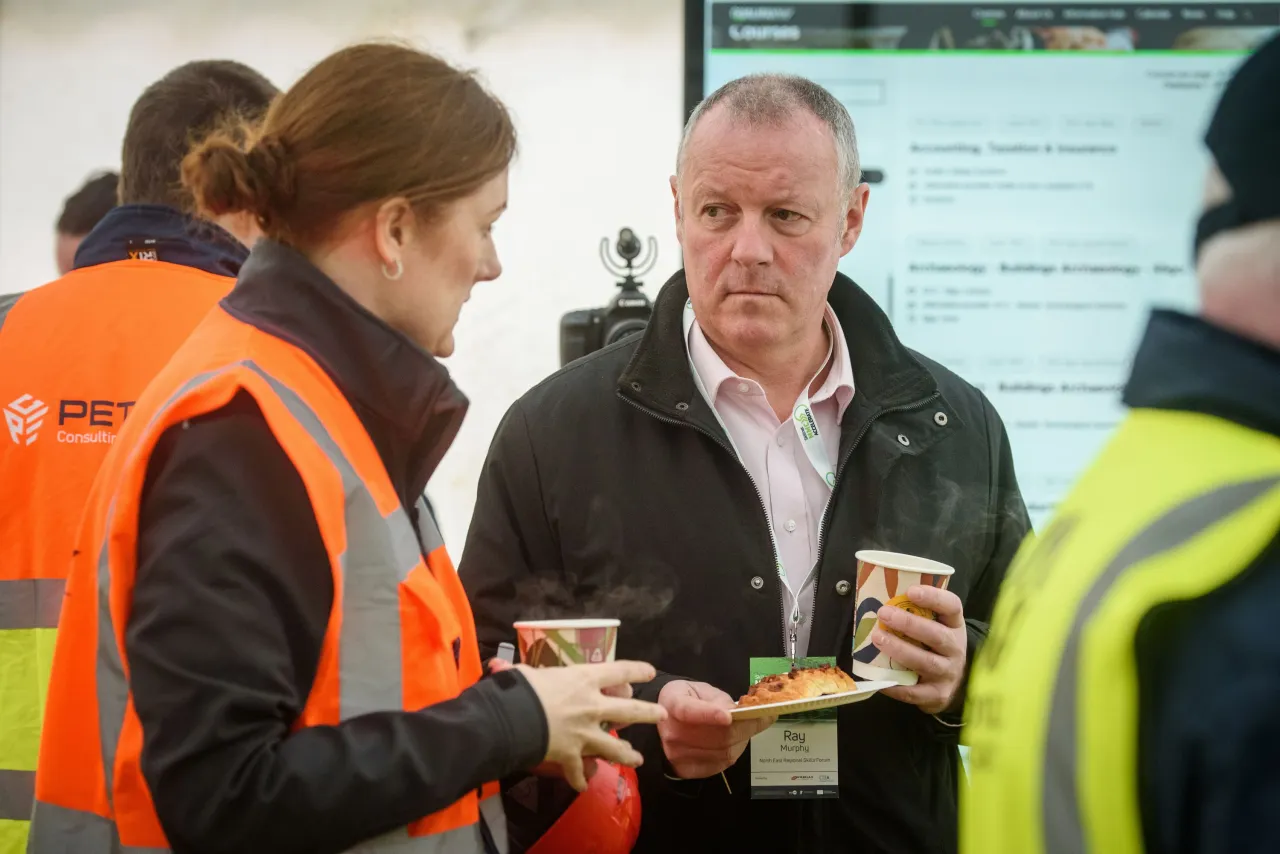 Two people in conversation at an outdoor event, one wearing an orange safety vest, both holding cups.