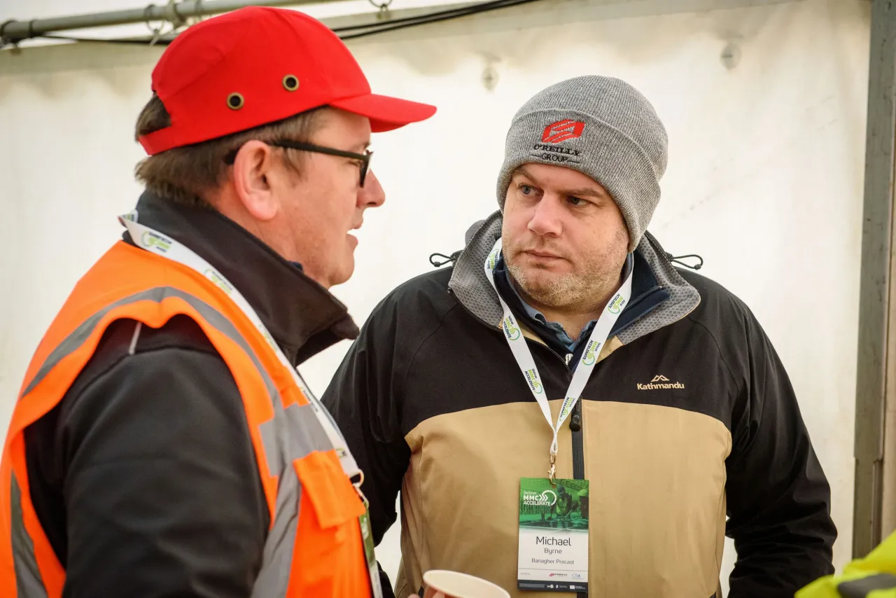 Two men in winter gear having a conversation outdoors, one wearing an orange safety vest and red cap.
