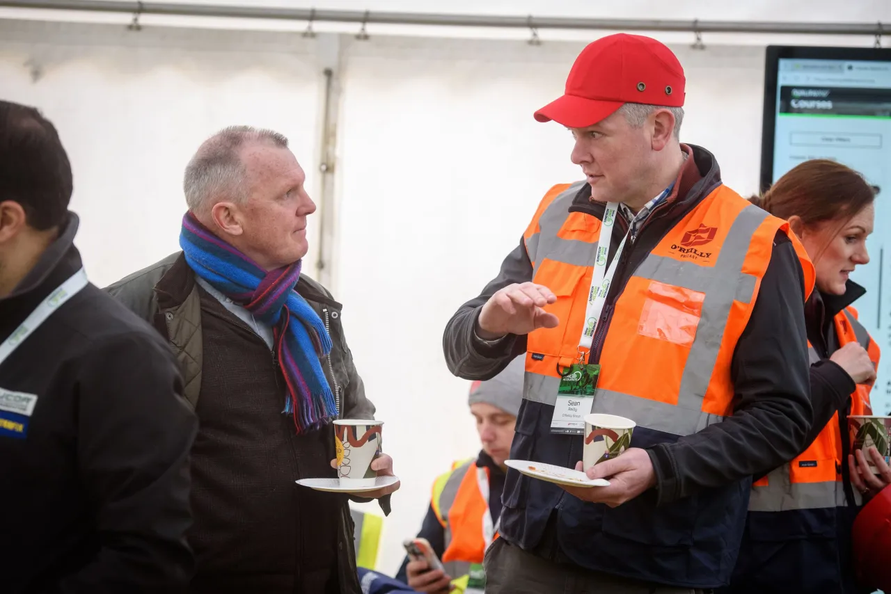 Two men talking at an outdoor event, one wearing an orange safety vest and red cap.
