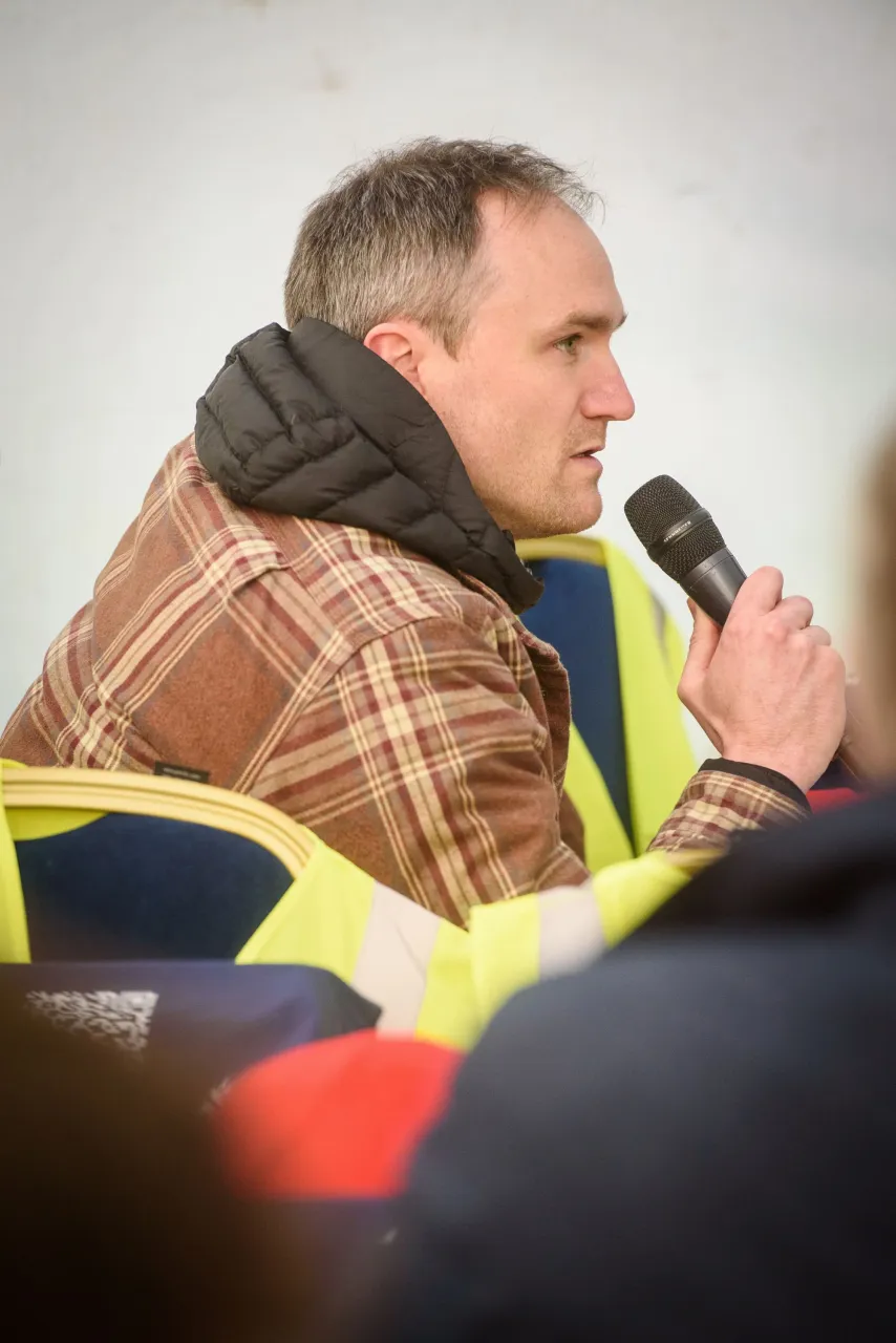 Man in a brown plaid jacket speaking into a microphone outdoors.
