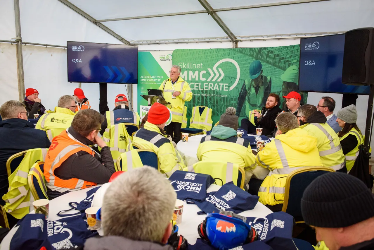 Group of workers in high-visibility vests attending a briefing under a tent at an outdoor event.