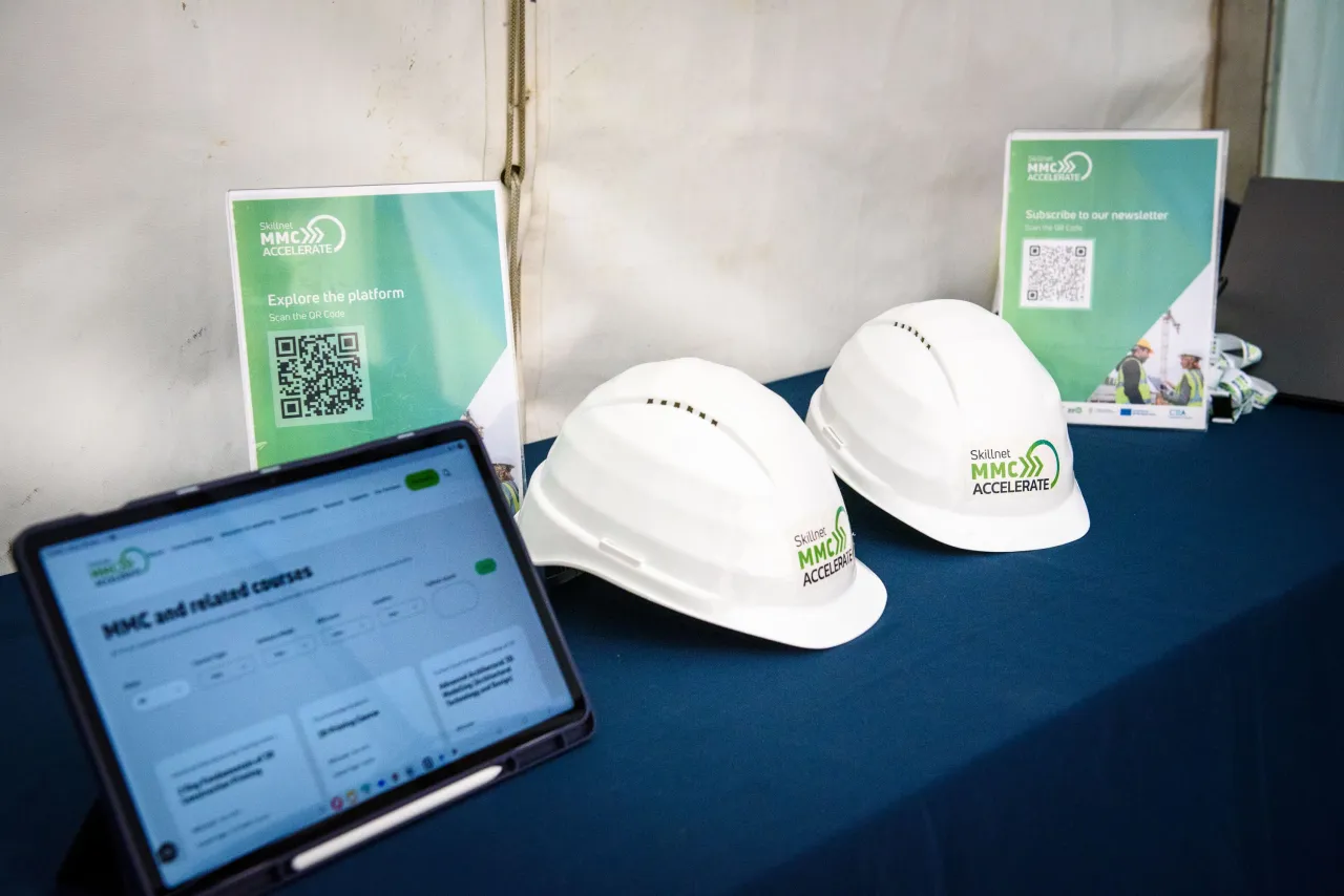 Two white hard hats with green logos displayed on a blue table beside a tablet device.