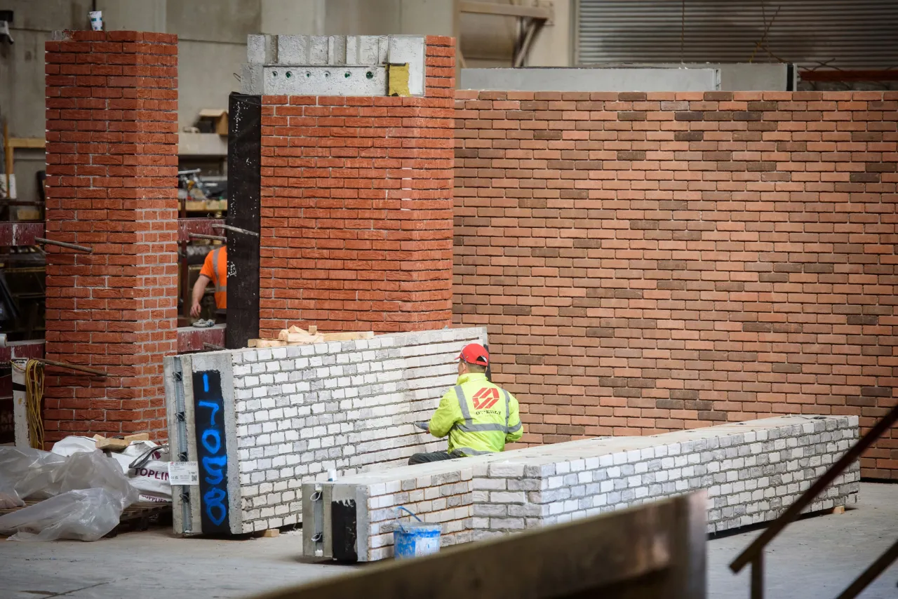 Construction worker in safety vest laying bricks on a partially built red brick wall indoors.