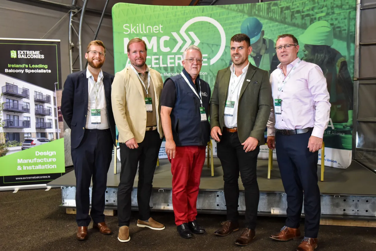 Five men posing together in front of a green event banner at a conference or trade show.