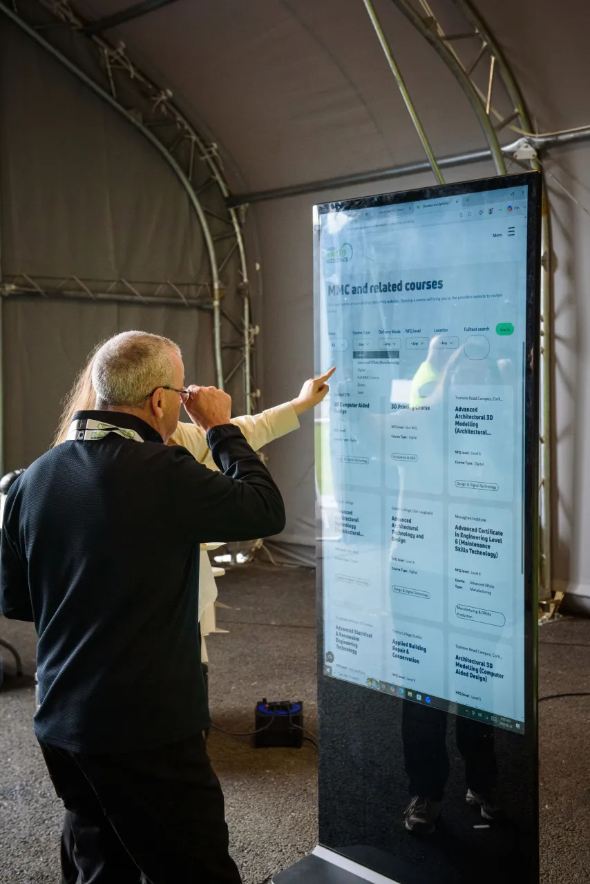 Man in black jacket pointing at a large digital display screen in an industrial setting.