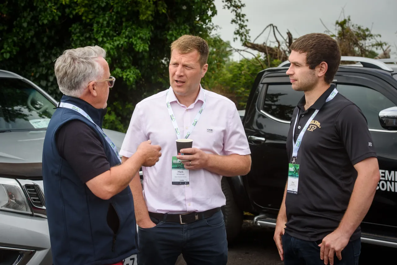 Three men in casual attire having a conversation outdoors near a dark SUV.