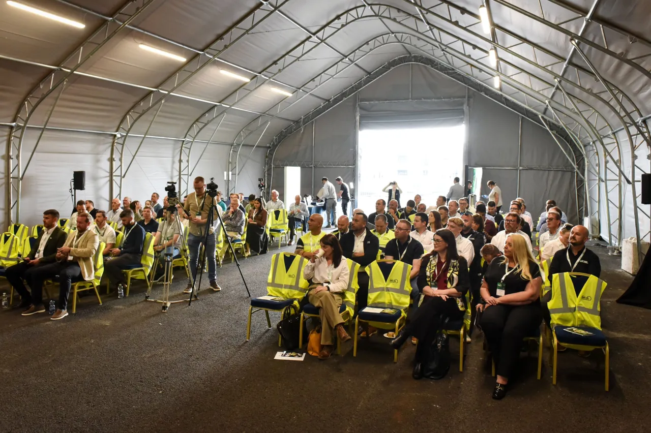 Large crowd seated in a temporary tent venue, some wearing yellow safety vests.