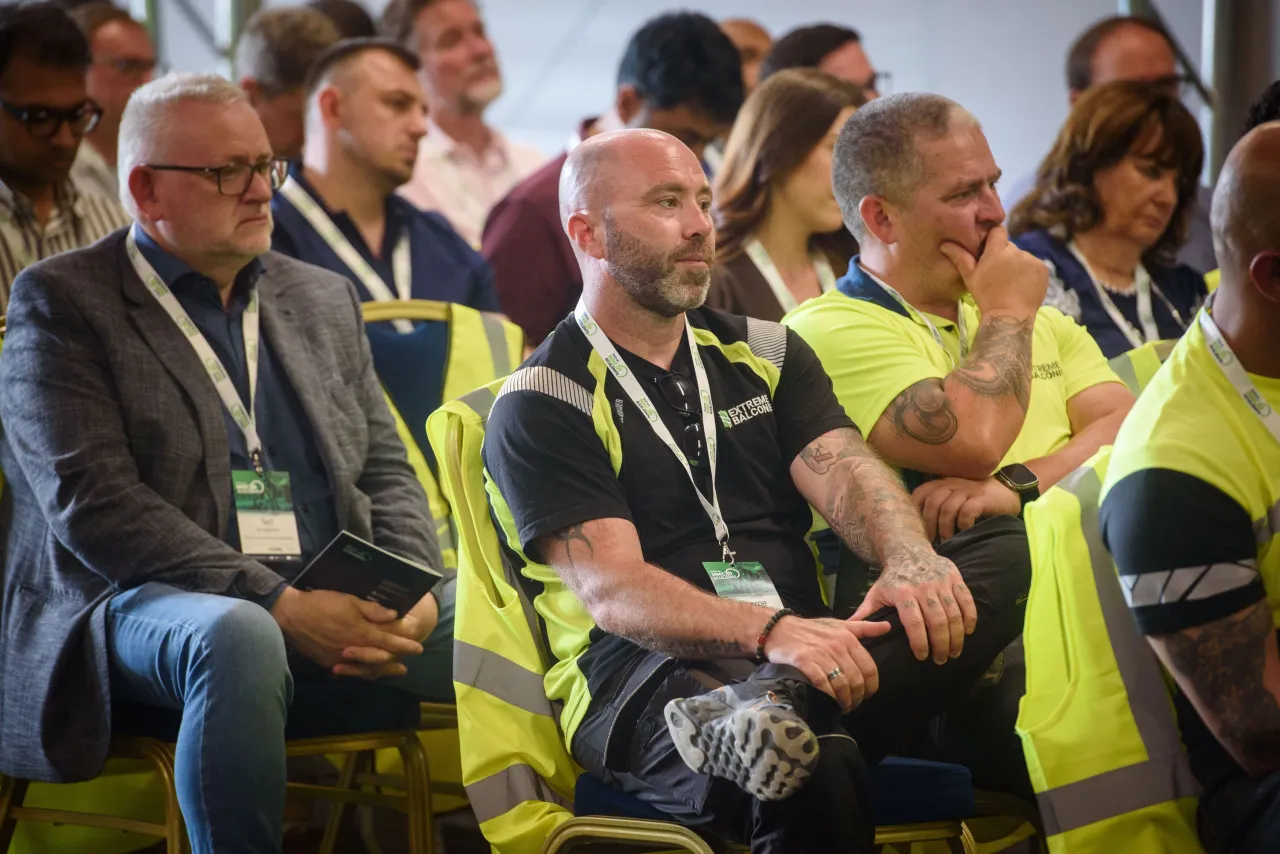 Audience members seated at an event, some wearing yellow safety vests and conference lanyards.
