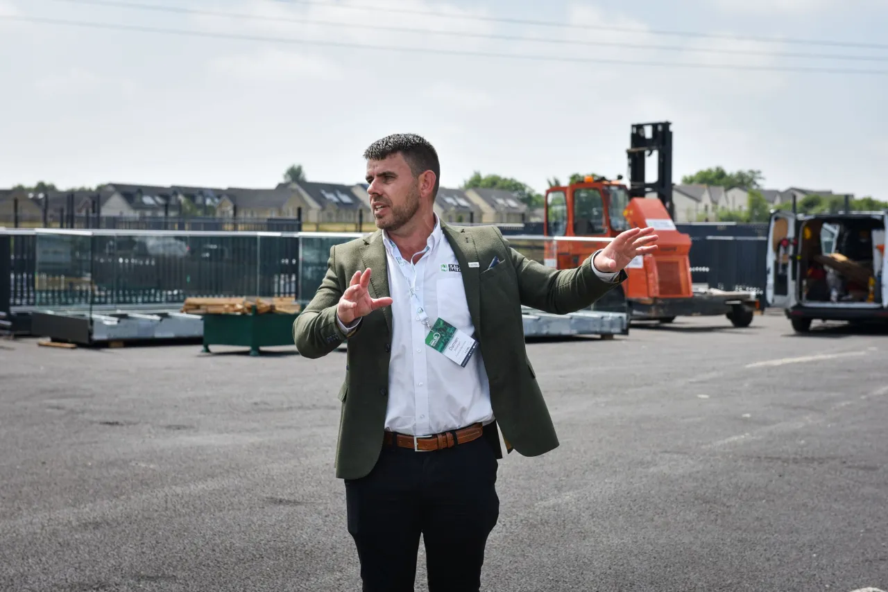 Man in a blazer gesturing outdoors at an industrial yard with a forklift in the background.
