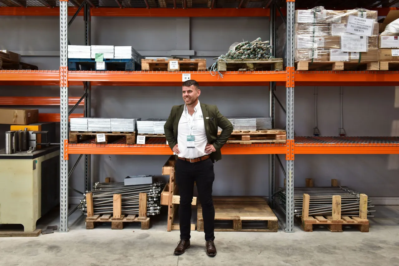 Man standing in a warehouse between orange metal shelving units holding a document.