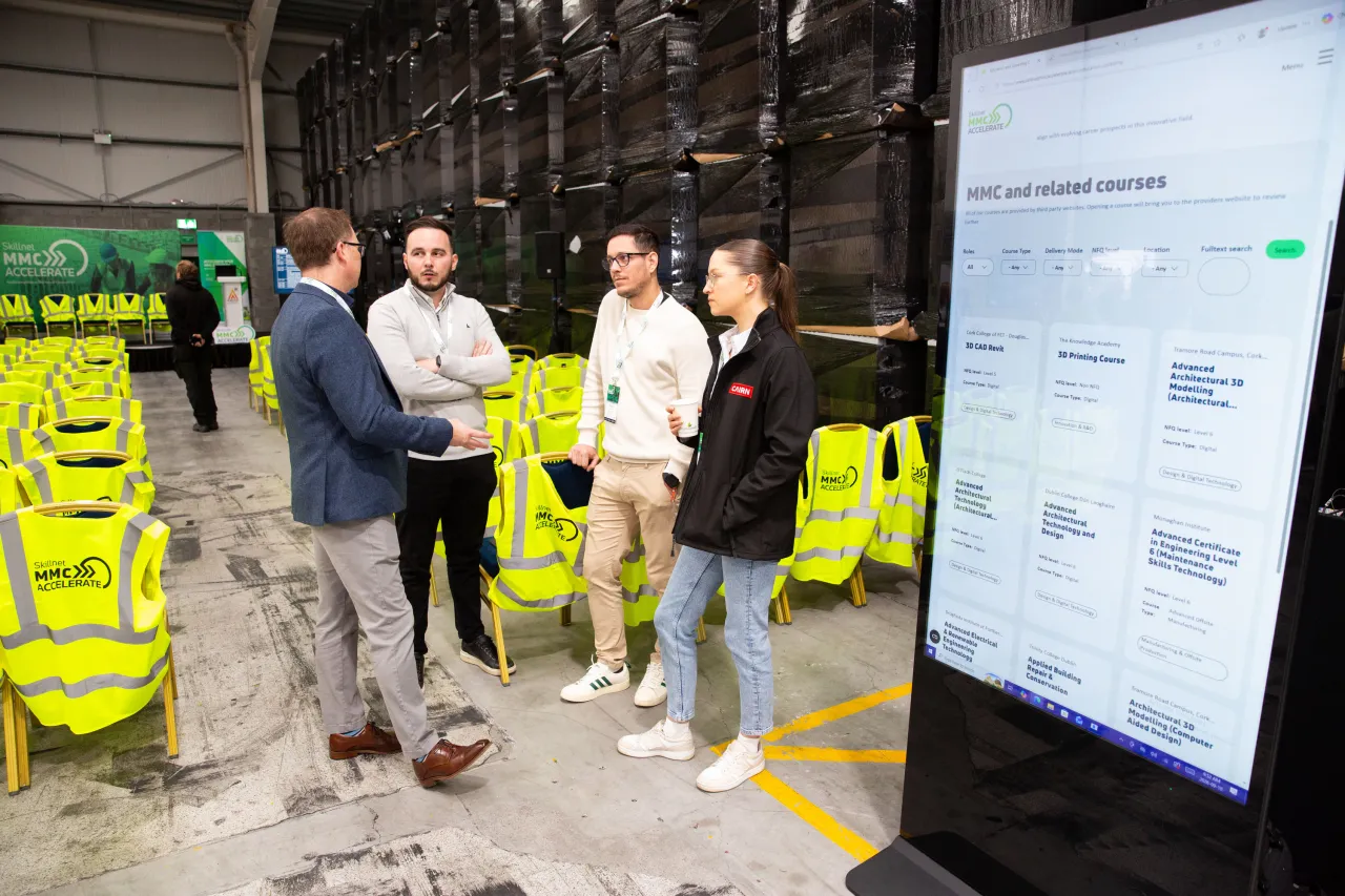 Four men conversing in an industrial warehouse surrounded by yellow safety vests and equipment.