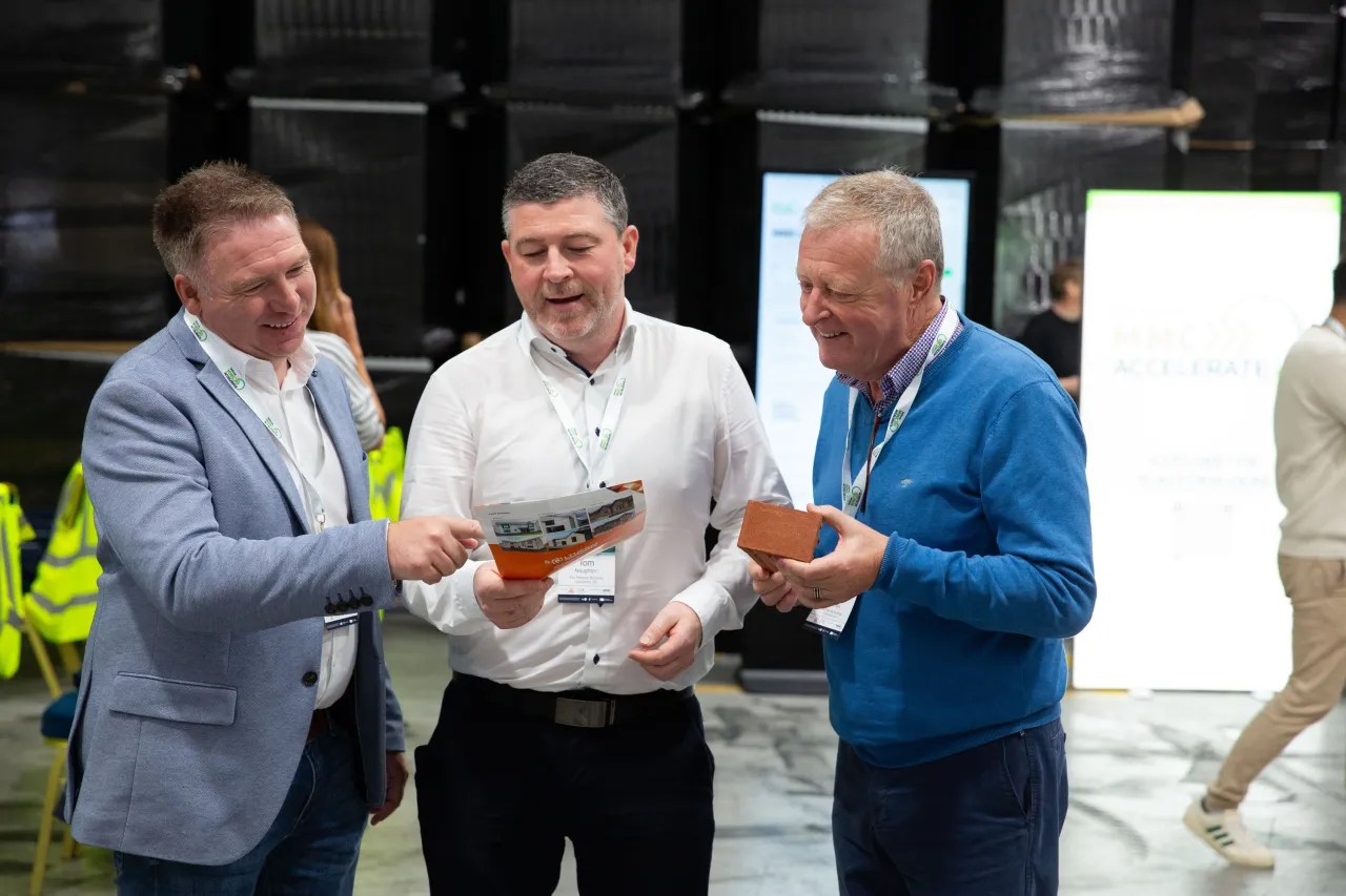 Three men smiling and examining an orange object together indoors.