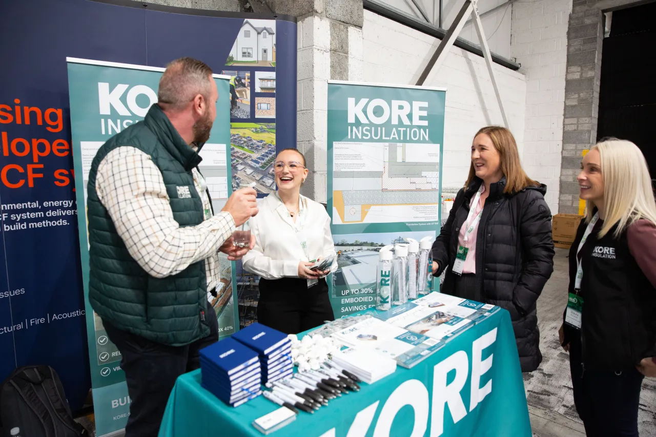 Three people conversing at a KORE exhibition stand during a business event.