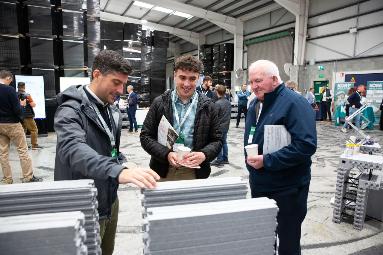 Three men examining tile samples at an indoor trade show or exhibition.