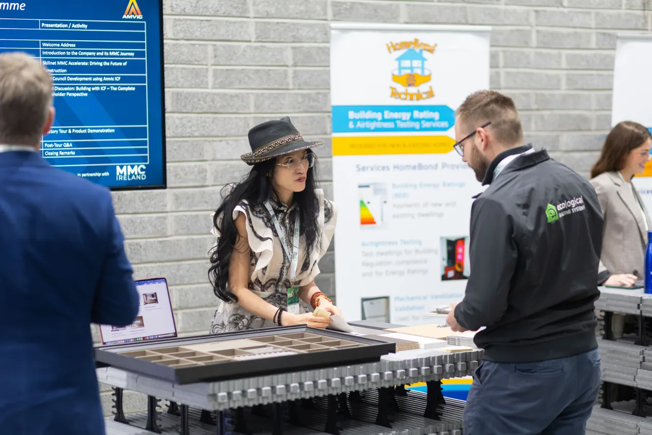 Woman in a hat at a trade show booth speaking with a man in a green jacket.