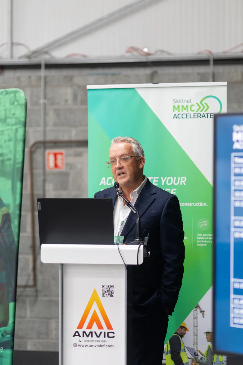 Man in a dark blazer speaking at a white podium with the AMVIC logo, beside a green banner.