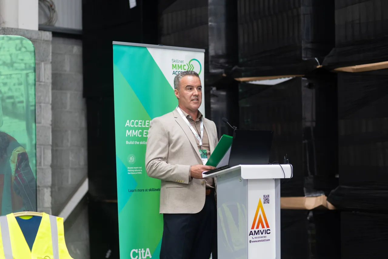Man in suit speaking at a podium with a green banner and warning sign nearby.