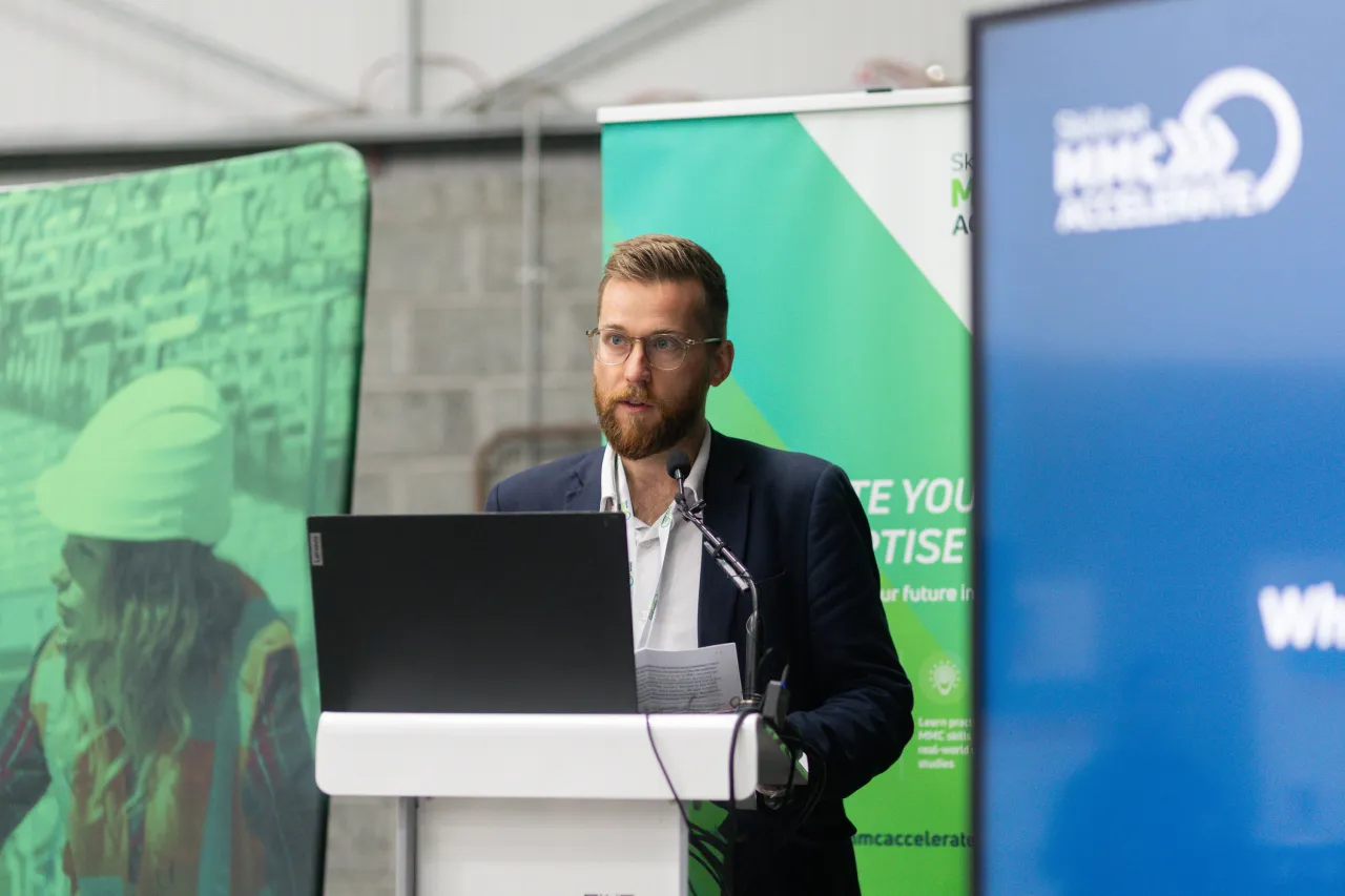 Man in blazer speaking at a podium with a green and blue banner backdrop outdoors.