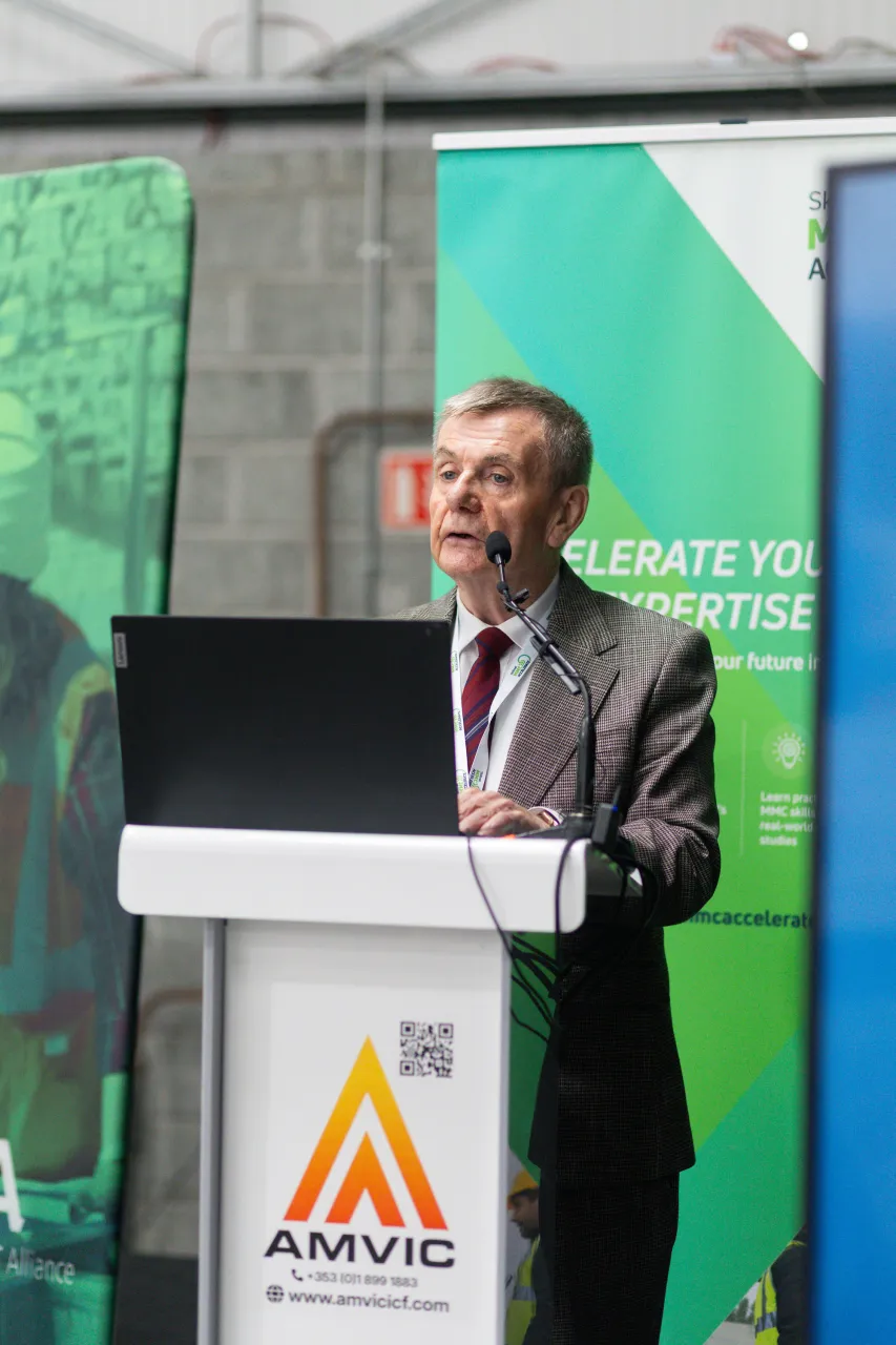 Older man in suit speaking at an AMVIC-branded podium during an outdoor event.