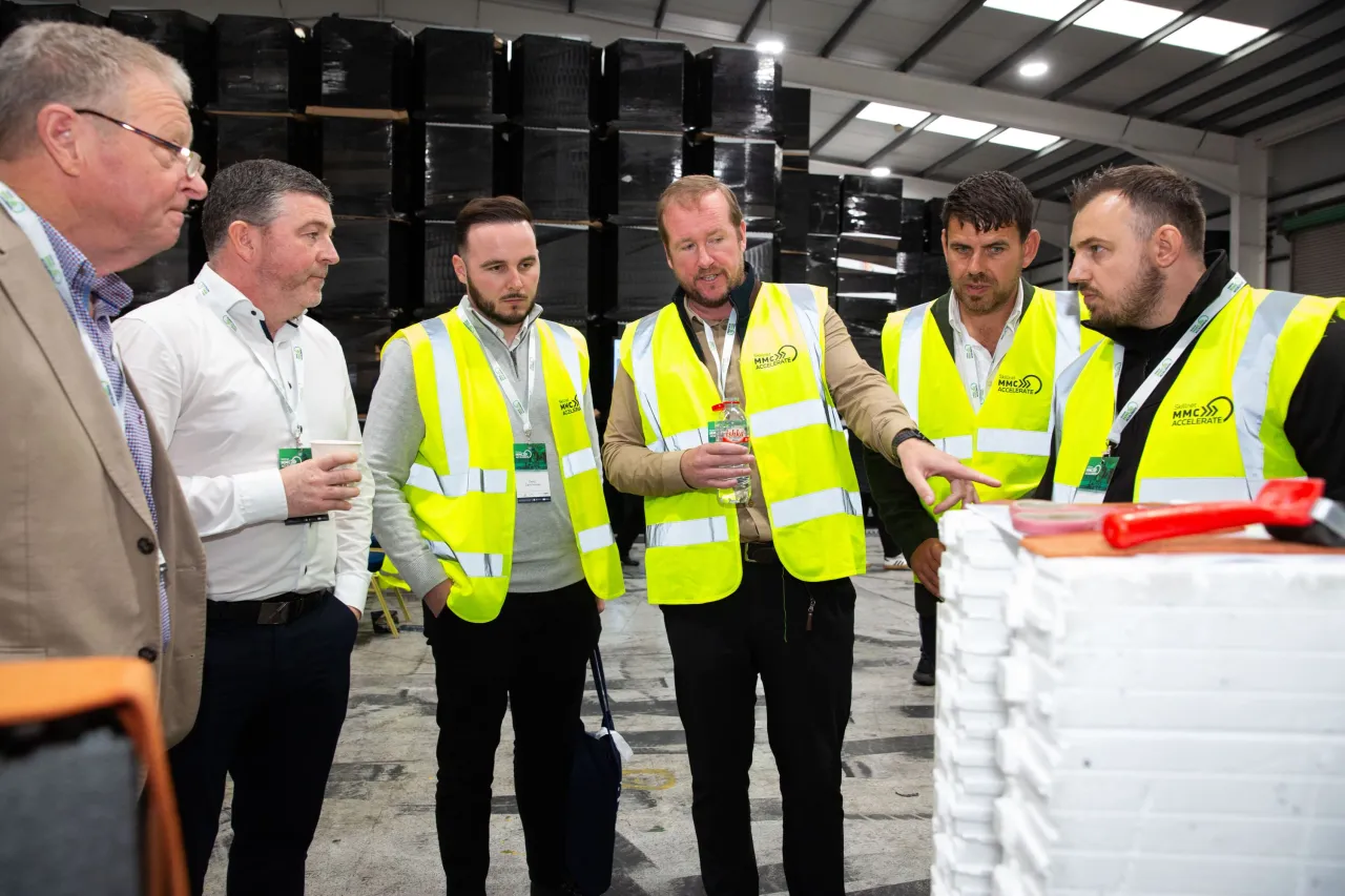 Five men in a warehouse, some wearing high-visibility vests, engaged in conversation among stacked materials.