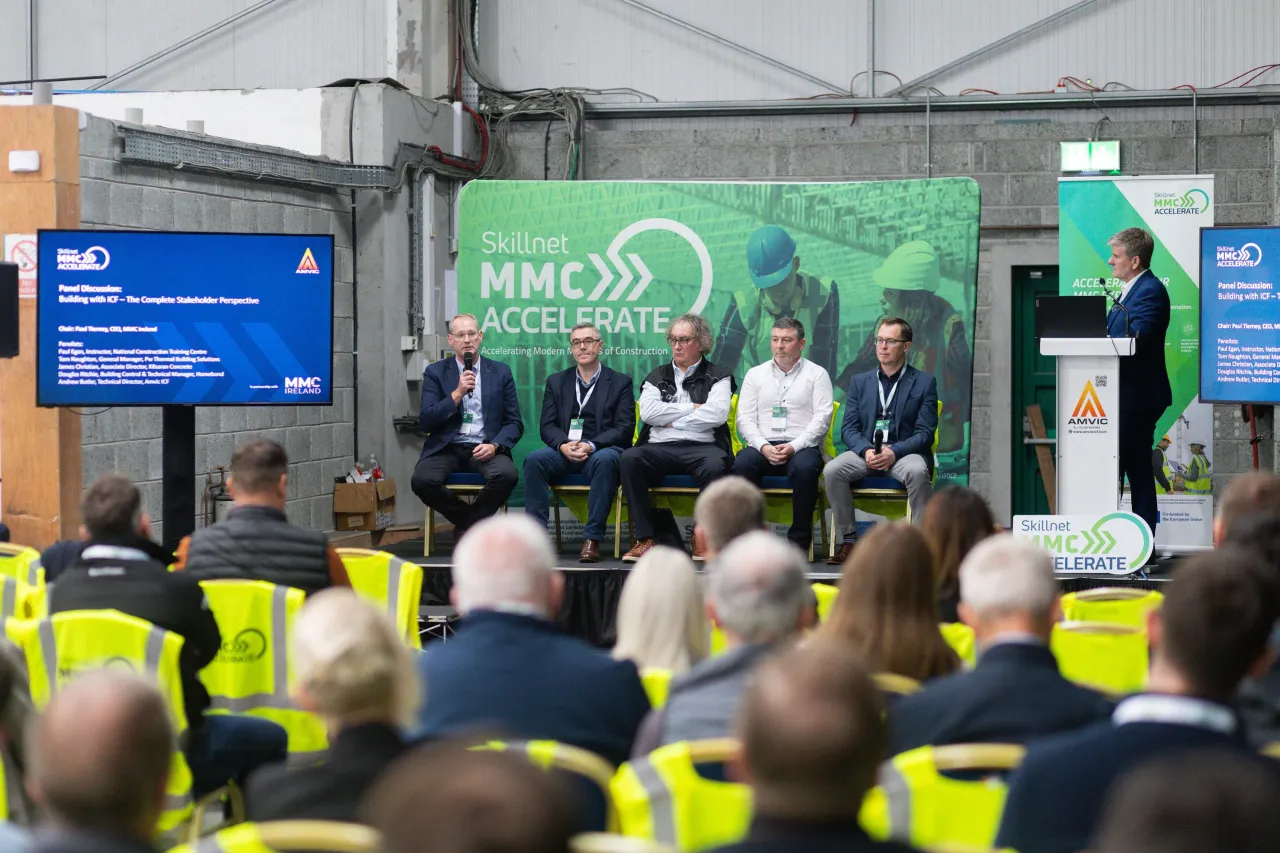 Panel discussion at an MHI event, speakers seated on stage before a crowd in safety vests.