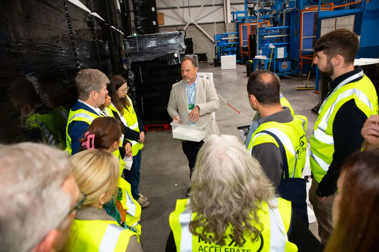 Group of workers in yellow safety vests listening to a speaker inside an industrial facility.