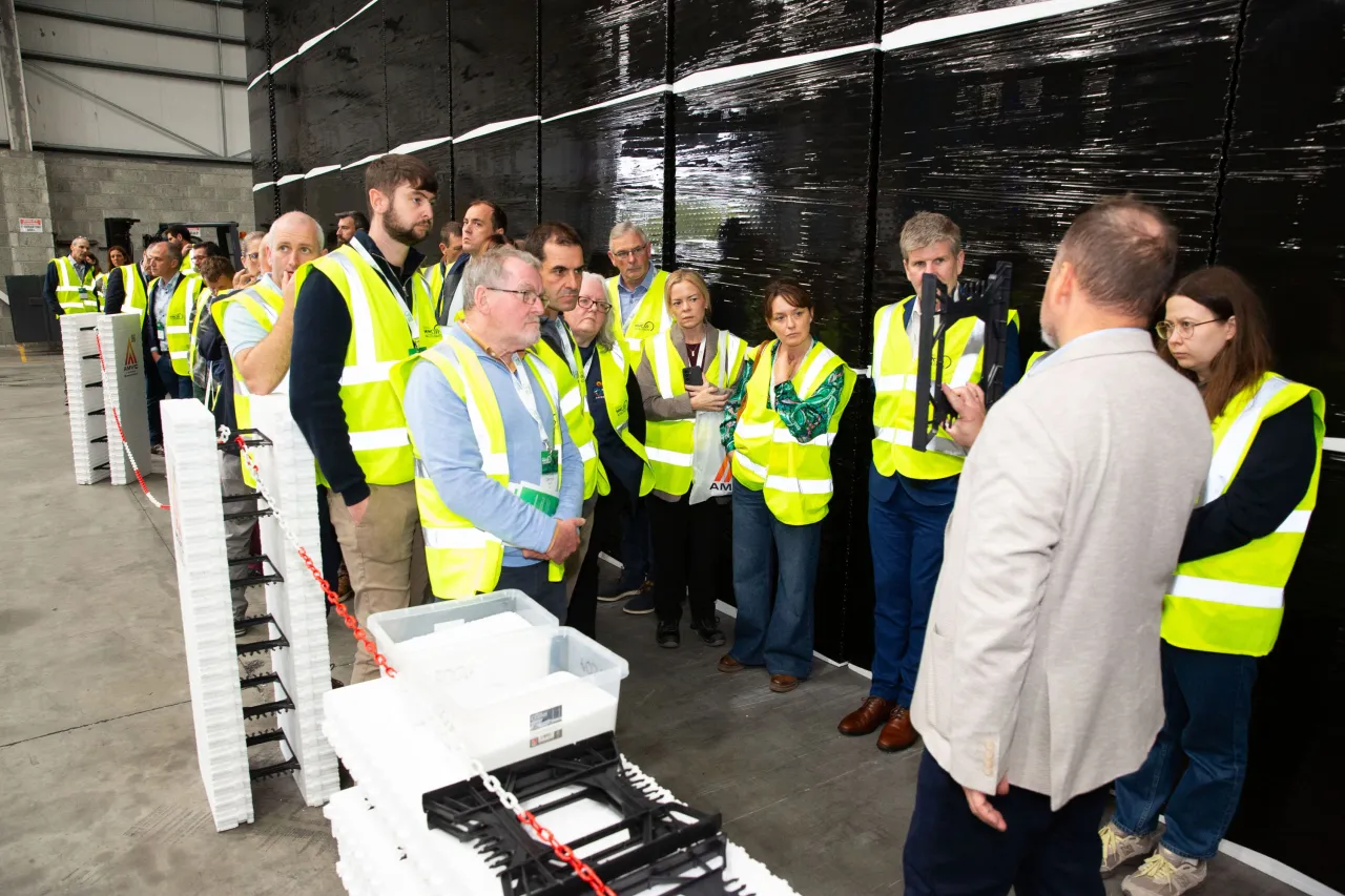 Group of people in yellow safety vests gathered around equipment in an industrial warehouse facility.