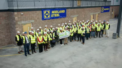 A group of people in high vis jackets gather outside a brick building with a sign for the company Midland Steel above.