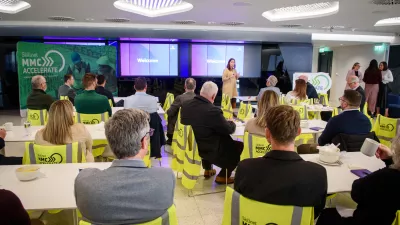 A group of people sit around tables watching a presentation from a female.