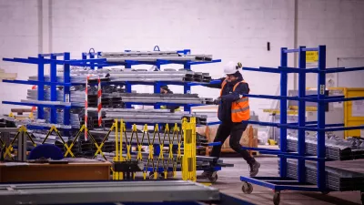 A male in a high vis jacket and hard hat manoeuvres a rack of steel frames on a trolley through a crowded manufacturing facility. 
