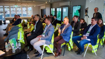 A group of people in business suits sit in rows listening to a presentation.