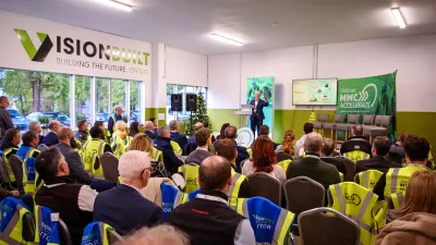 A group of people sit watching a presentation by a person in a blue suit standing behind a podium.