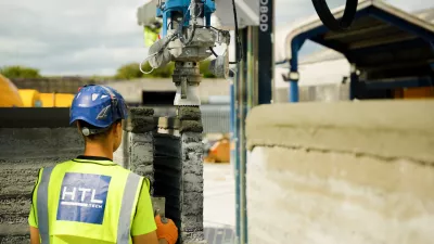 Person with a high-vis jacket and blue helmet watches as a 3D printer lays concrete on a wall.