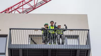 A group of three people in hard hats, protective goggles, and high vis jackets smiling on a metal balcony.