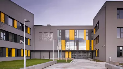 A modern grey school building with yellow and grey panels, surrounding a grass courtyard.
