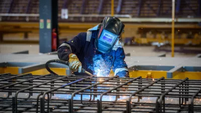 A person in protective clothing and a welding helmet uses a blowtorch to weld steel bars in a manufacturing facility.