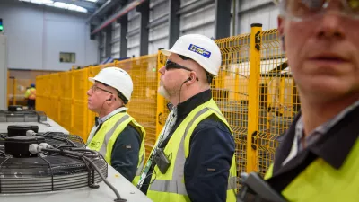 Three people in high-vis vests and hard hats observing machinery inside a construction factory.